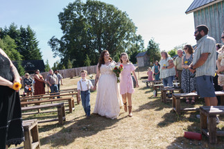 Bride walks down outdoor aisle with two children, guests watching.