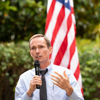 Man speaking at microphone, American flag backdrop