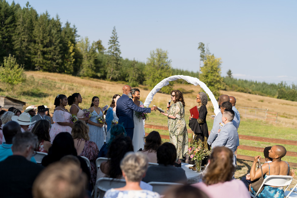 Outdoor wedding ceremony, celebrant officiating couple under white arch. Guests watching.