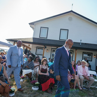 Two men in suits walking past seated guests at an intimate farm wedding.