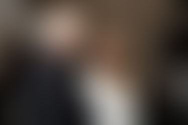 Man with white hair and glasses talks to red-haired woman in white dress.