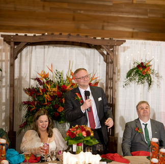 Man gives wedding speech at reception table with smiling guests.