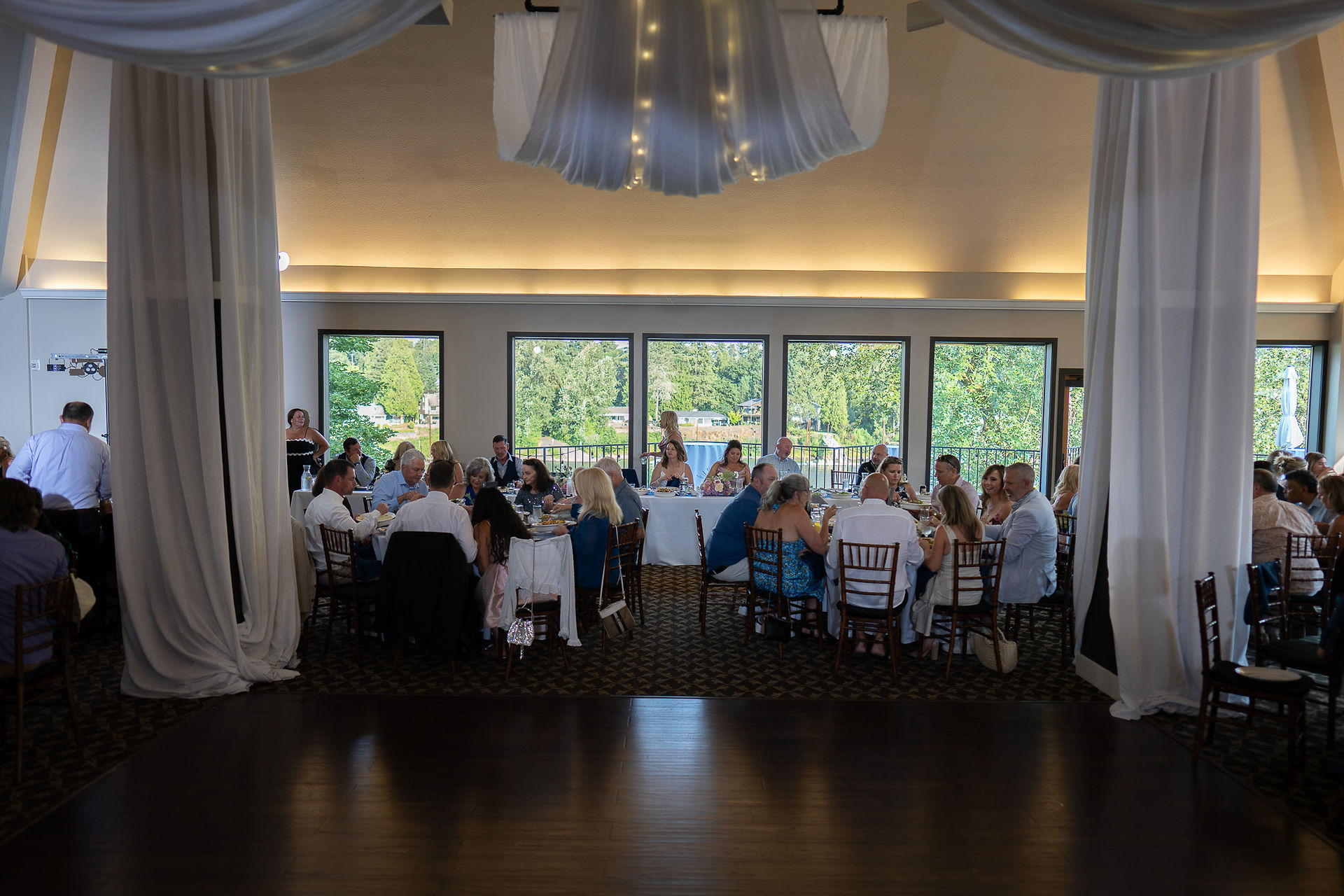 Wedding reception guests seated at tables, elegant indoor venue with lake view.