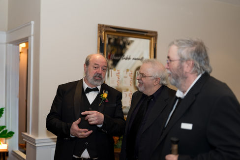 Bearded man in tuxedo gestures while talking with two men.