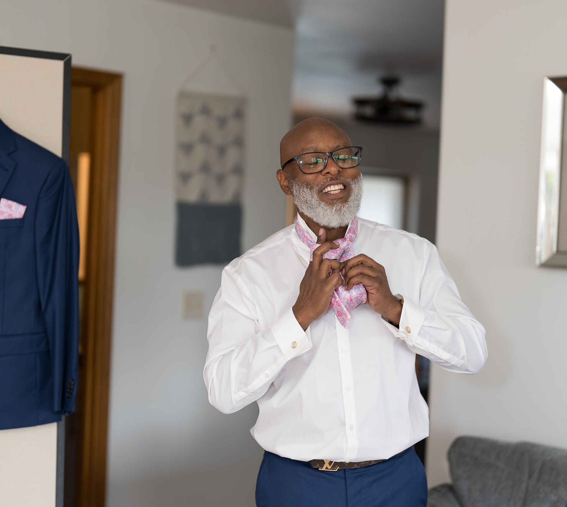 Smiling bearded man wearing glasses ties a pink tie indoors.