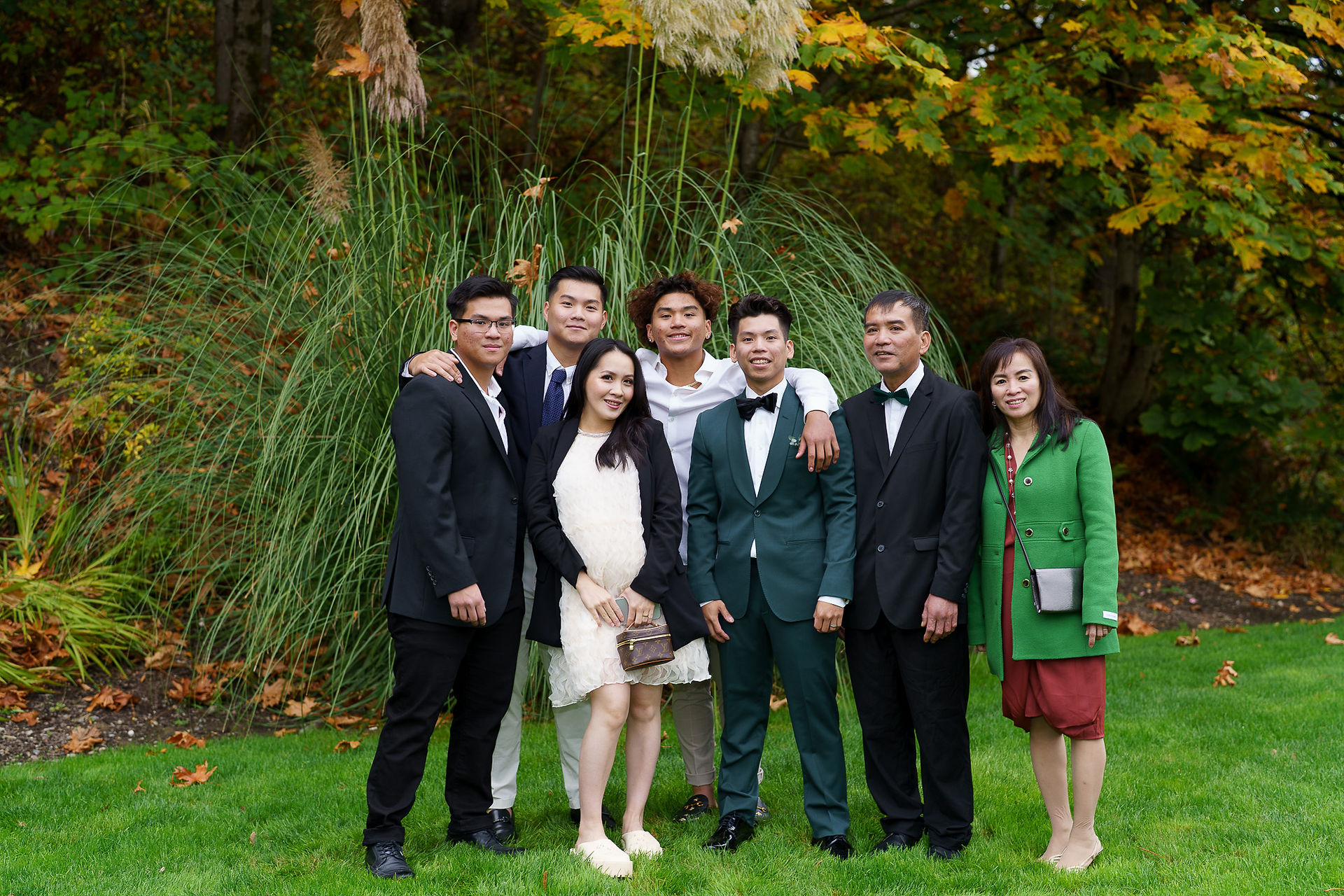 Seven formally dressed guests posing outdoors amidst lush green foliage.