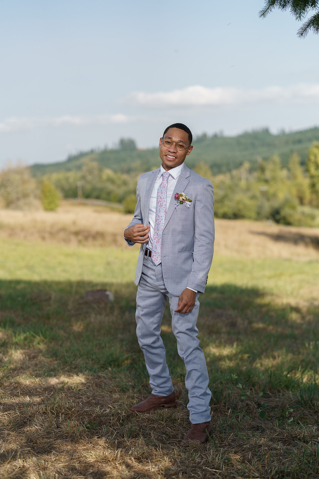 Man in light grey suit adjusting tie outdoors, smiling.
