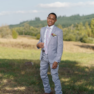Man in light grey suit adjusting tie outdoors, smiling.