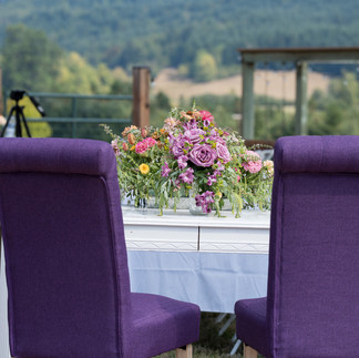 Elegant outdoor table with floral centerpiece, purple chairs, and hills.