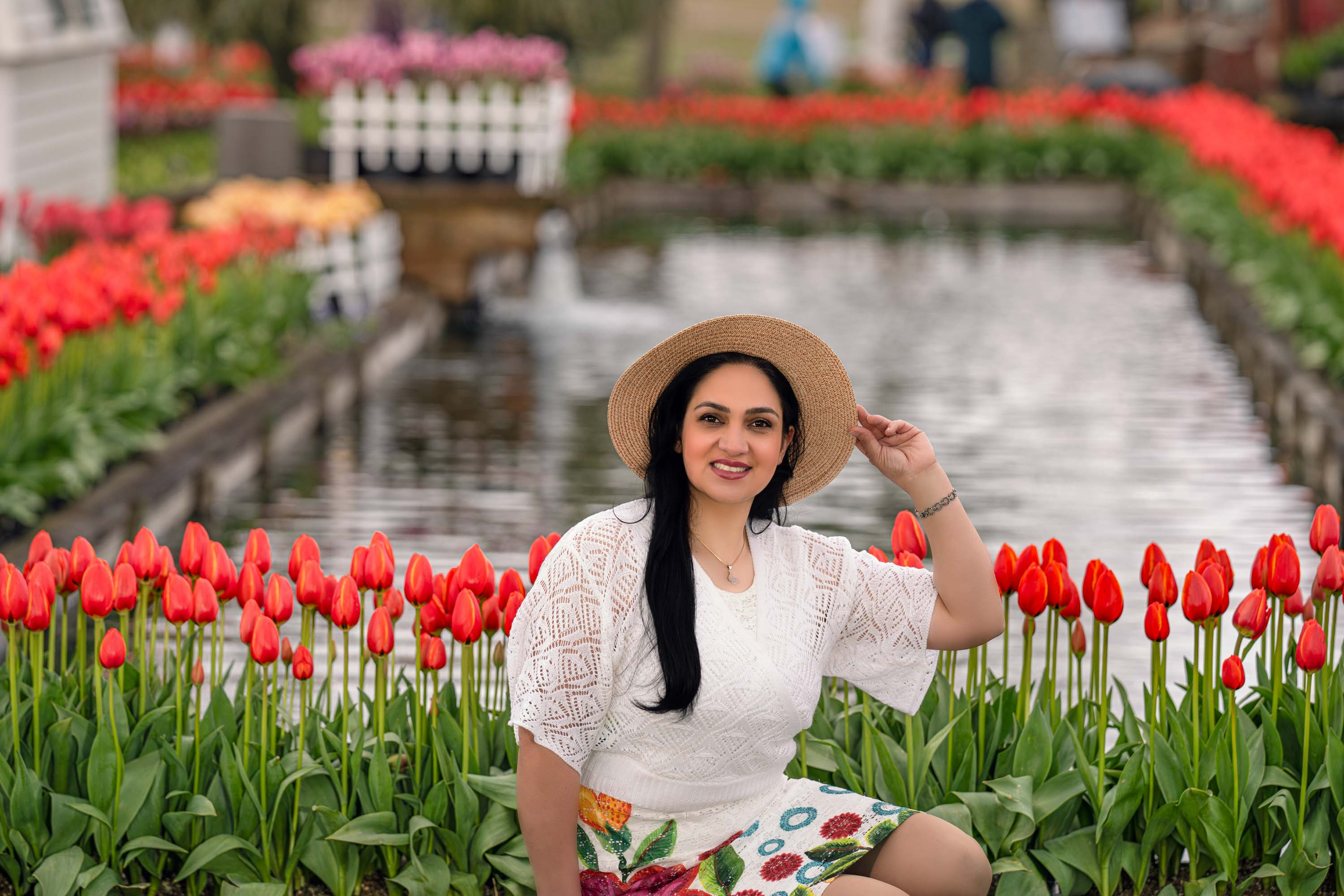 Smiling woman in hat amidst vibrant red tulips, pond, at tulip festival 2026.