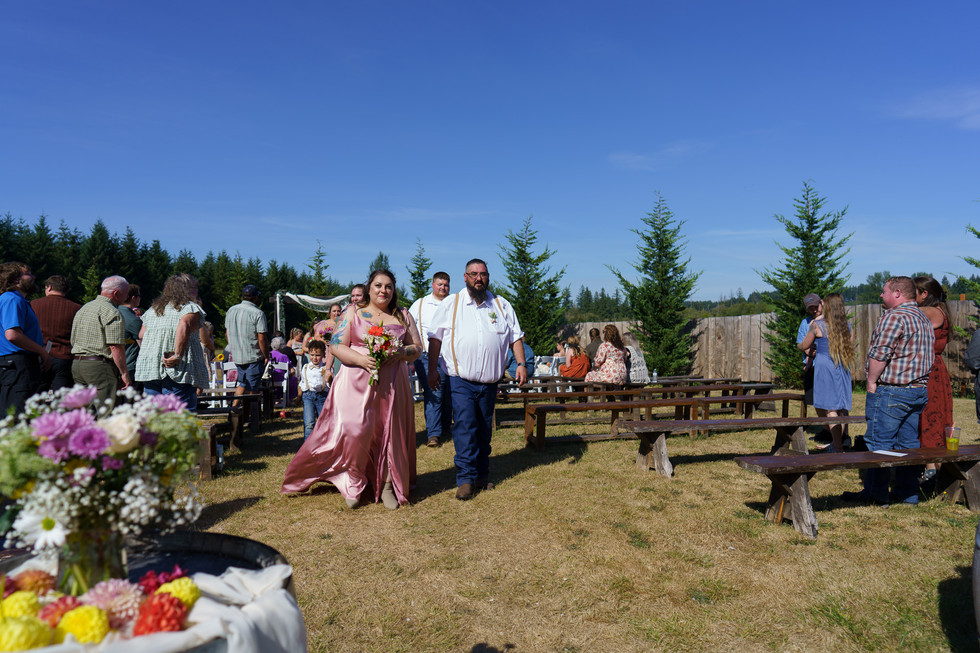 Woman in pink gown and man walk down aisle at outdoor wedding.