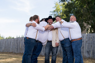 Groomsmen embracing a smiling groom in a cowboy hat outdoors on a sunny day.