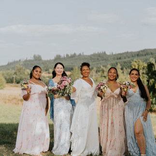 Five diverse women in pastel floral dresses holding bouquets outdoors.