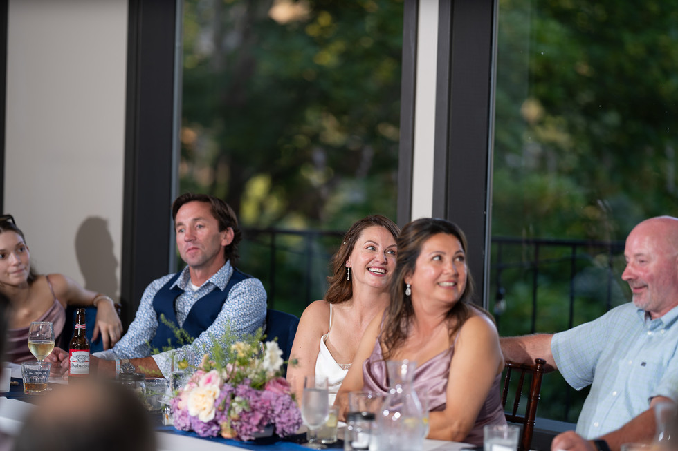 Guests smiling, attentive at table with floral centerpiece, scenic view.