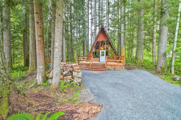 A-frame cabin in a lush forest setting, surrounded by tall trees and a gravel path. Logs are stacked beside the cabin, evoking a serene retreat.