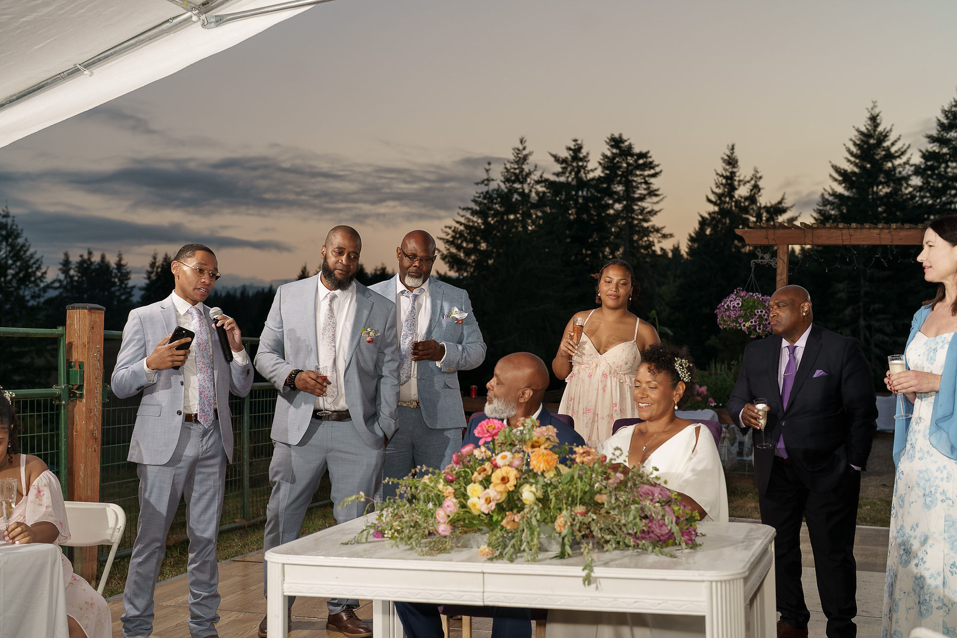 Man giving speech at outdoor wedding reception with guests listening.