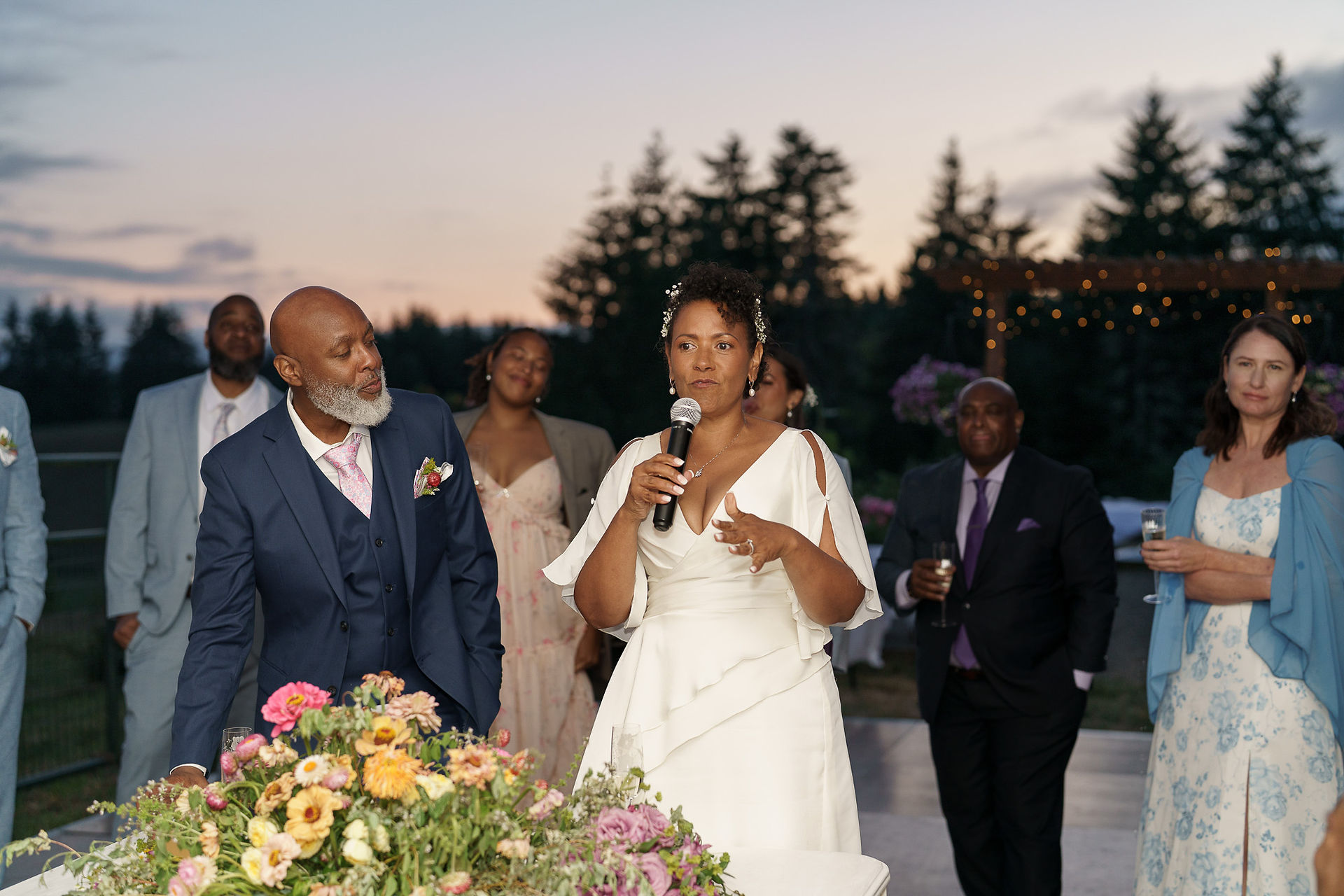 Bride in white dress speaks into microphone at outdoor wedding reception.
