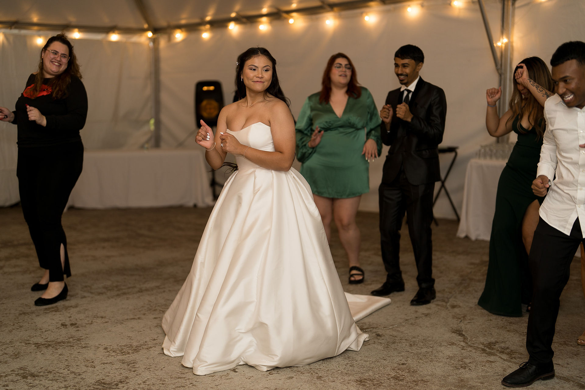 Happy bride dancing at wedding reception as groom watches nearby.