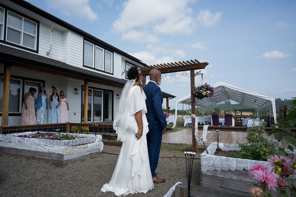 Bride and groom stand outside farmhouse, looking towards outdoor wedding tent.