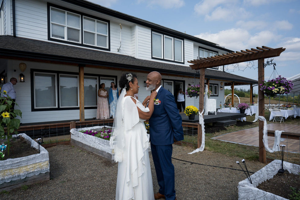 Bride adjusts groom's tie lovingly outside farmhouse. Intimate Farm Wedding.