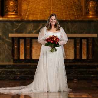 Smiling bride in white gown holding red bouquet inside ornate chapel.