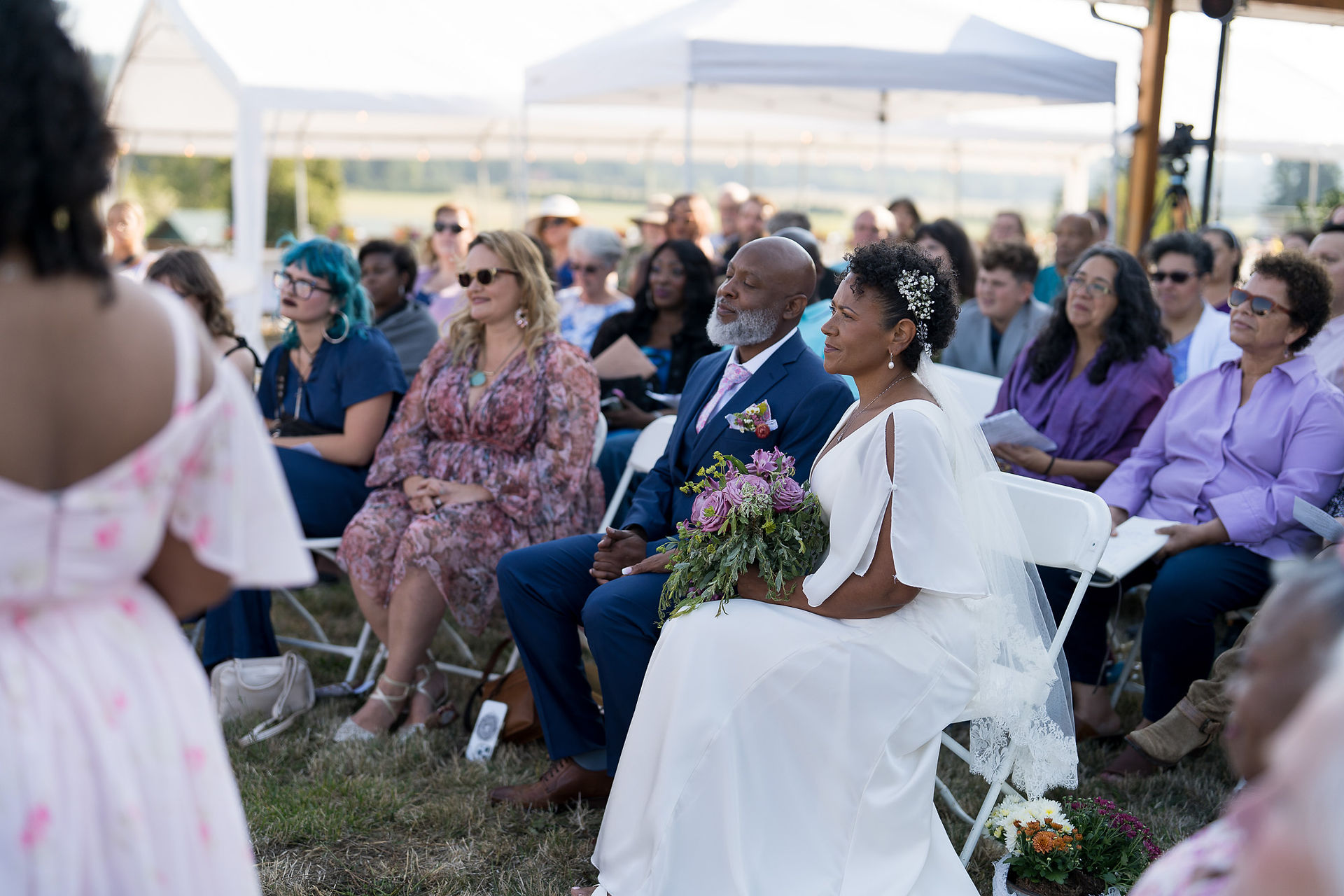 Wedding guests, including bride and groom, seated at an outdoor ceremony.