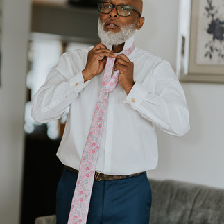 Man with white beard tying a pink floral tie indoors.