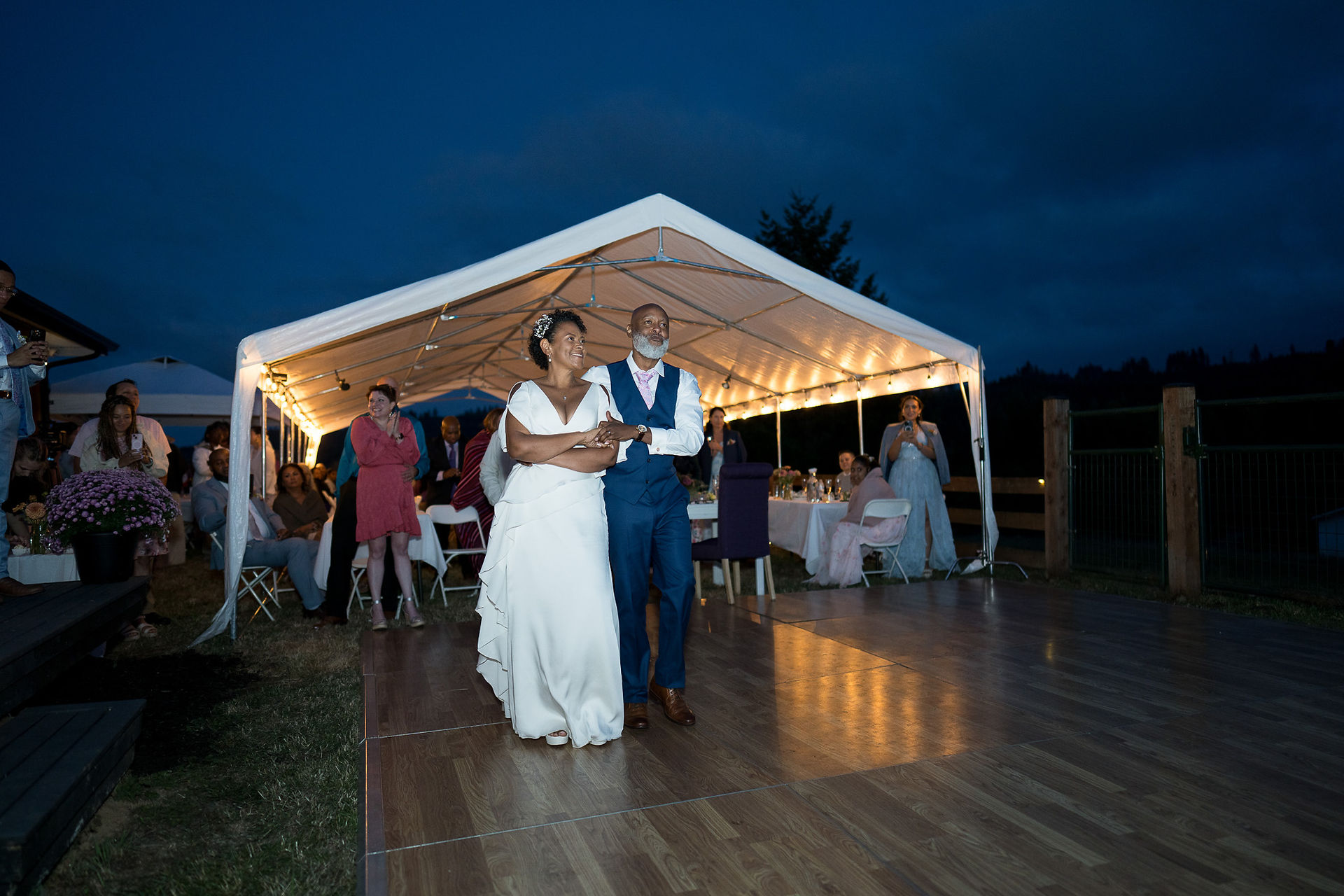 Married couple walks on dance floor under illuminated tent at intimate farm wedding.