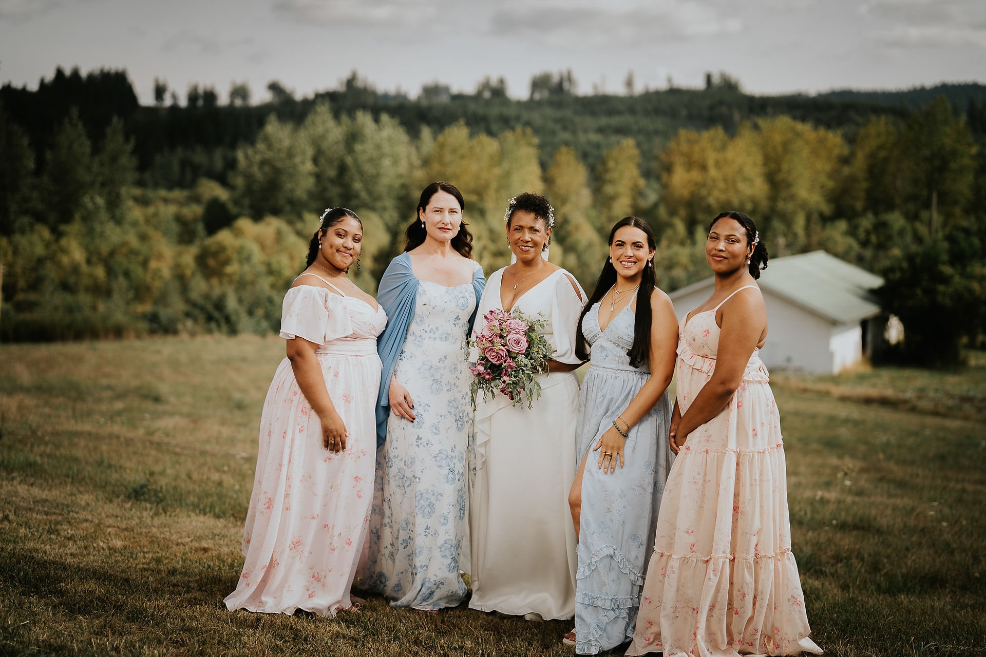 Bride with four bridesmaids in elegant dresses holding a flower bouquet outdoors.