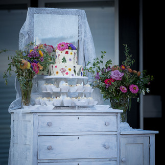 Rustic dresser with wedding cake, cupcakes, and colorful flowers outdoors.