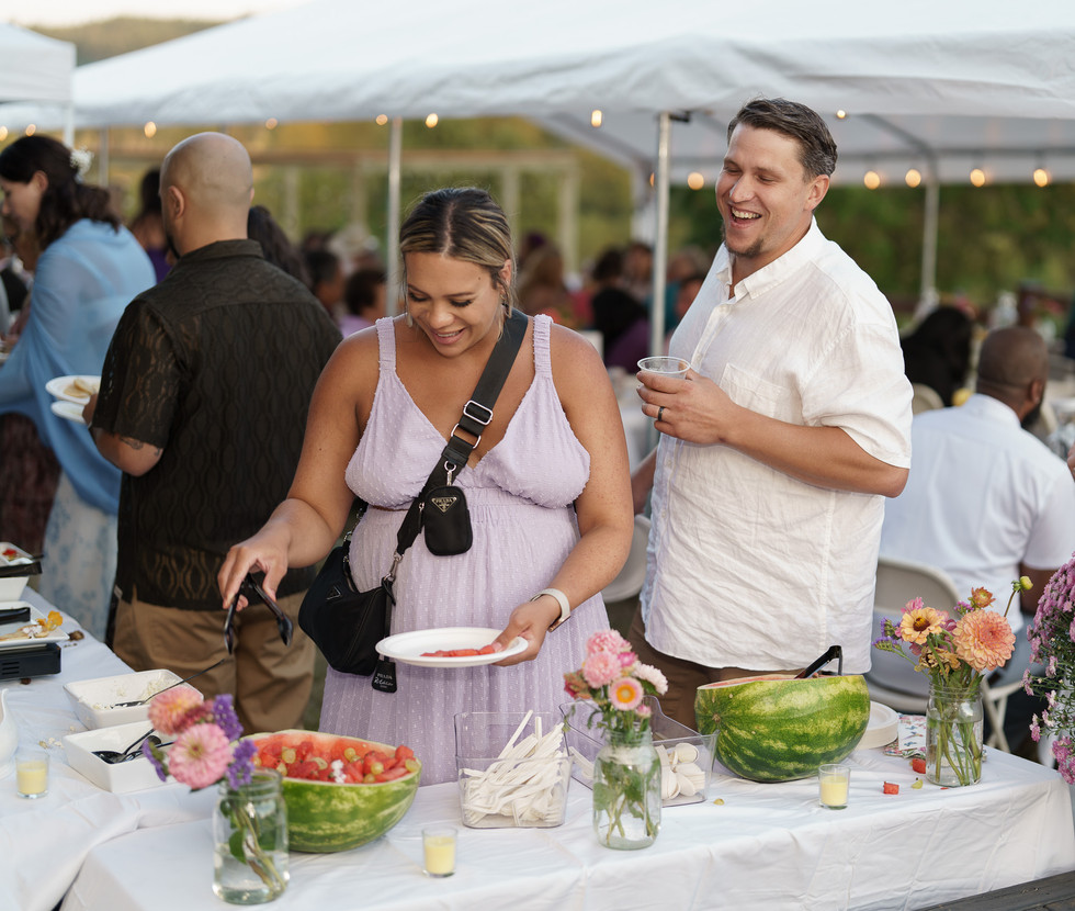 Woman serves watermelon from outdoor buffet as man laughs beside her.