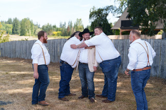 Groomsmen hug groom in cowboy hat and suspenders on wedding day.