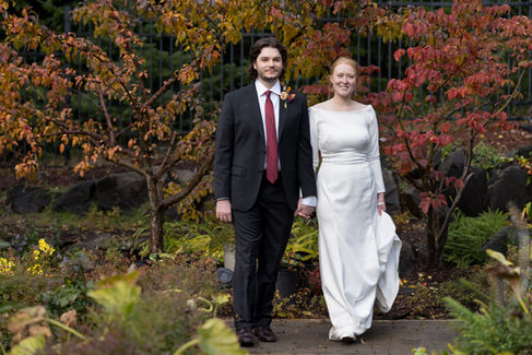 Happy groom and bride holding hands among colorful autumn trees.