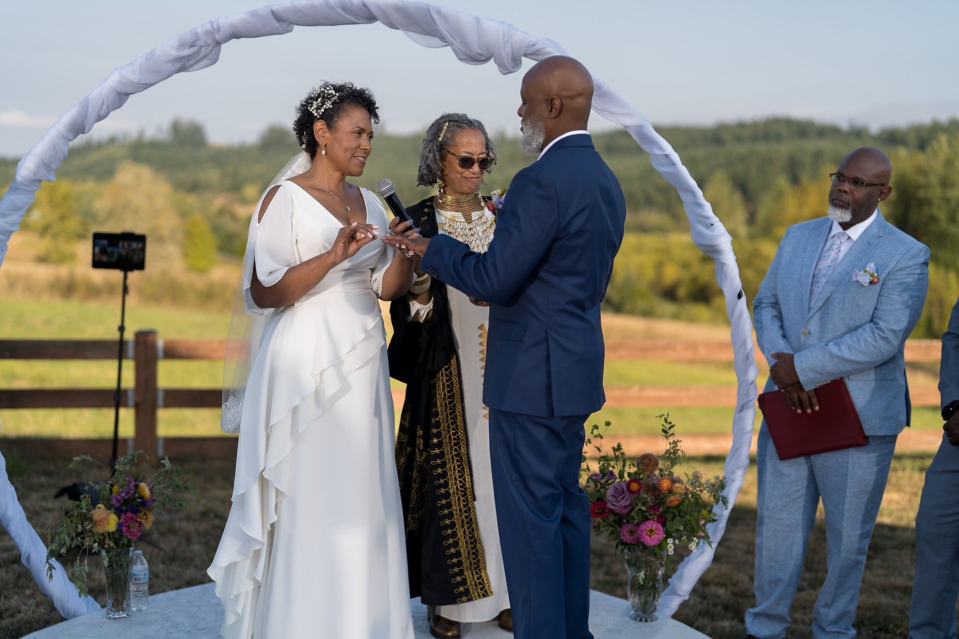 Bride, groom, officiant at intimate farm wedding ceremony under white arch.