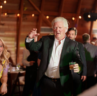 White-haired man in suit playfully dancing with a drink at reception.
