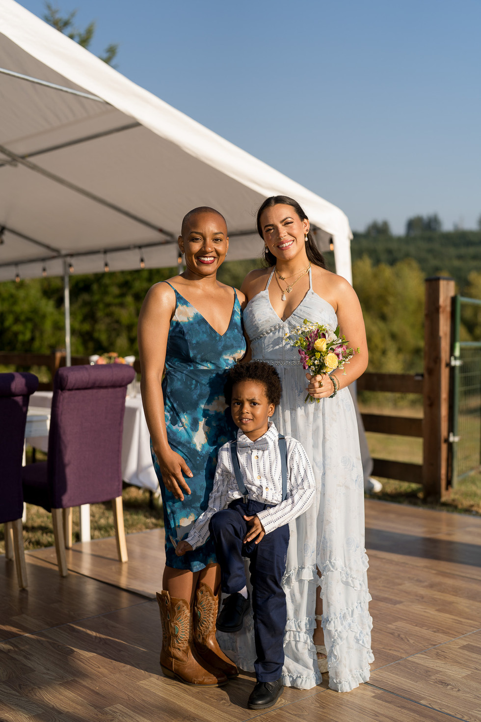 Two smiling women, boy with bouquet under outdoor tent.