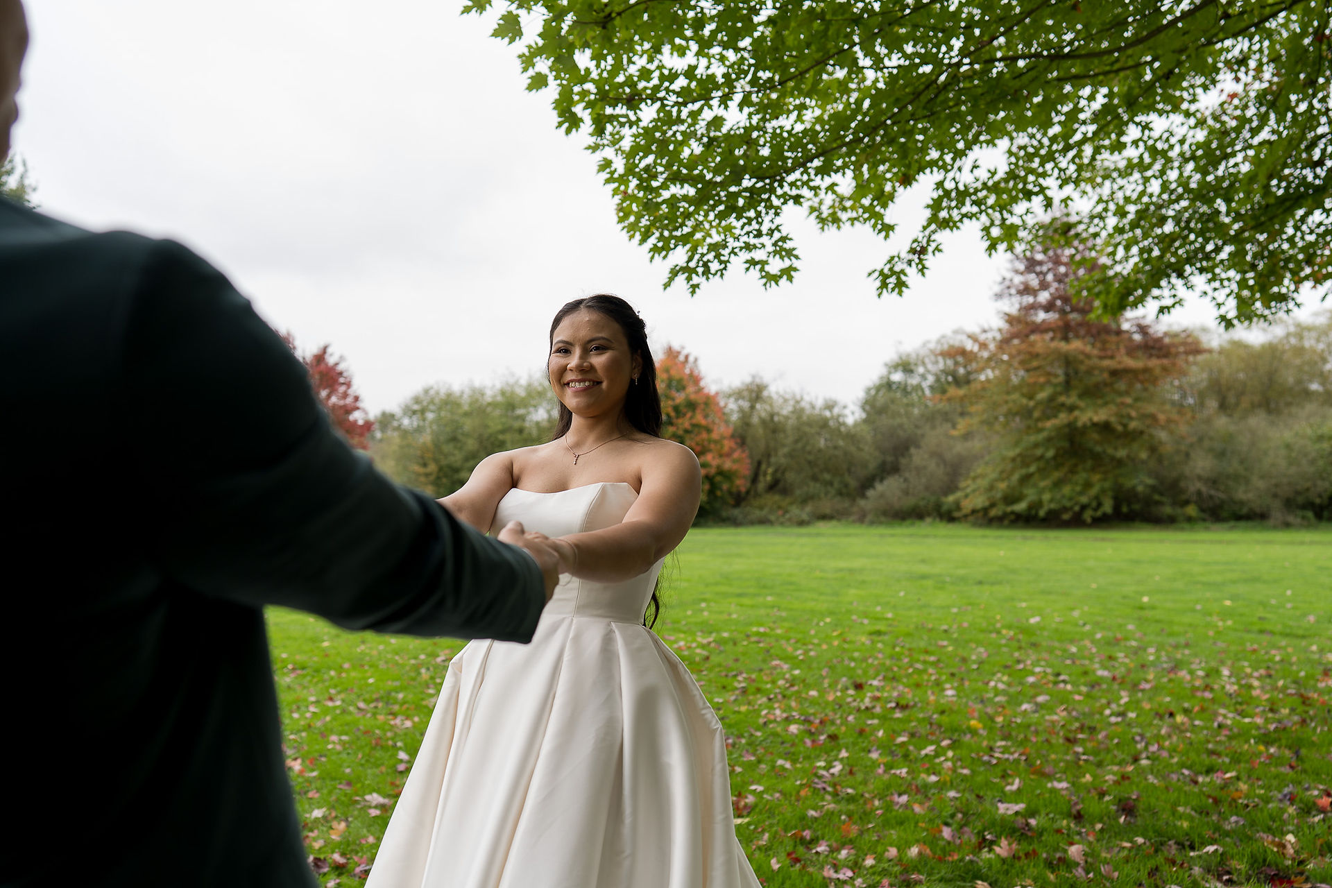 Smiling bride in white gown holding hand in lush green field.
