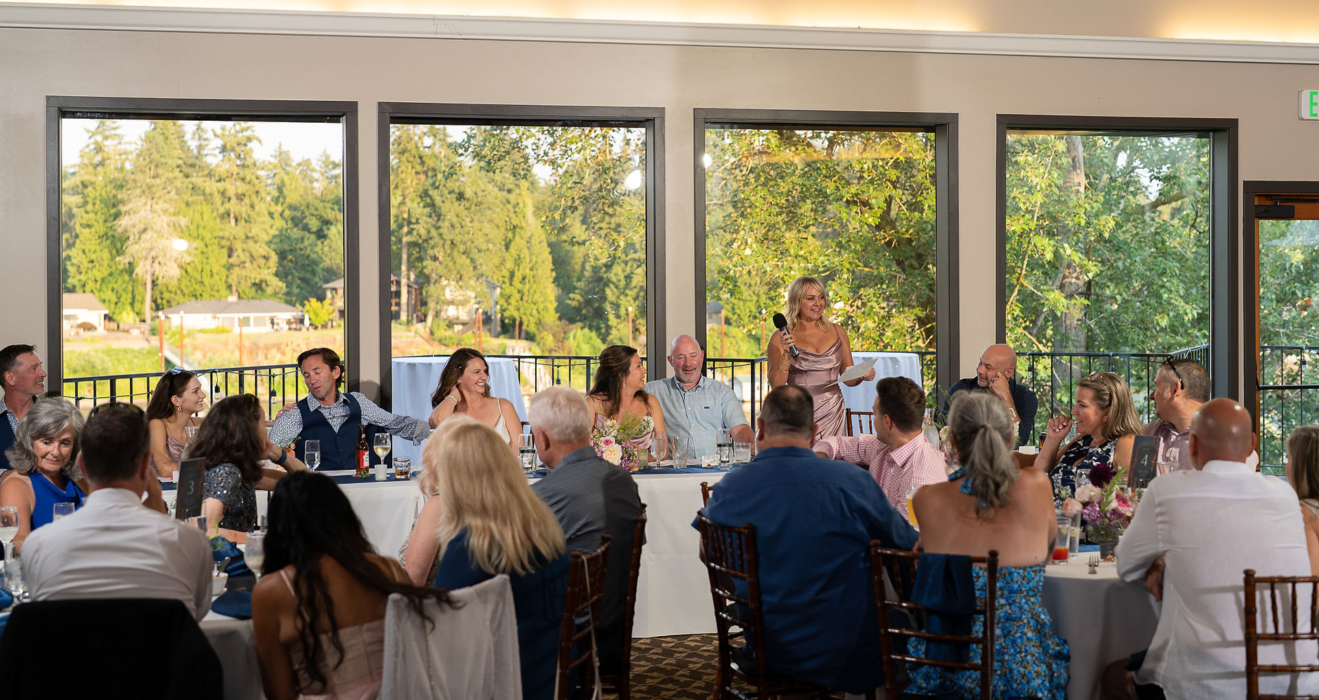 Woman speaking at a reception table with guests and an outdoor view.
