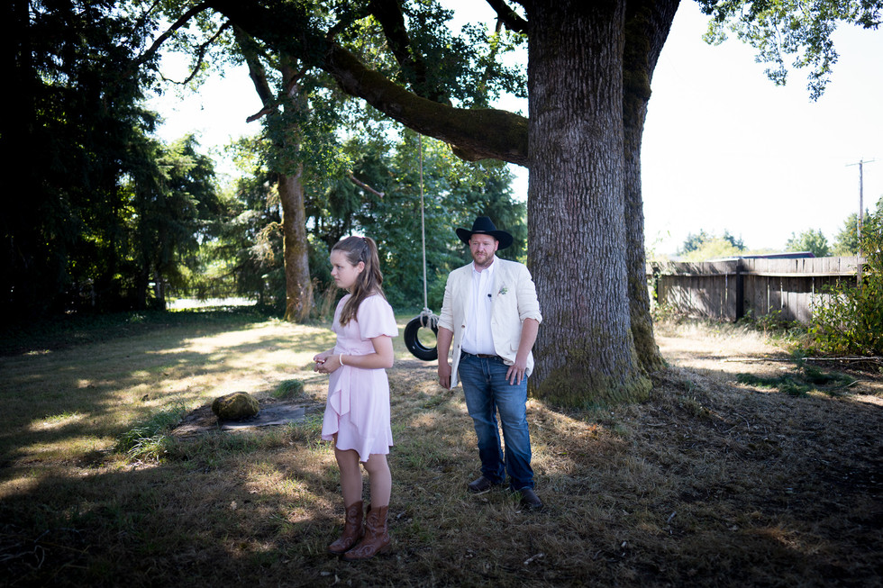 Girl in pink dress, man in cowboy hat, under large trees.