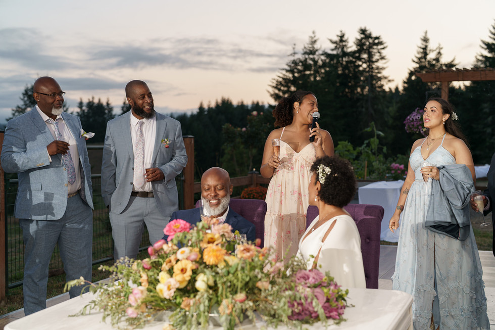 Woman speaks into microphone, guests smile at outdoor wedding reception.
