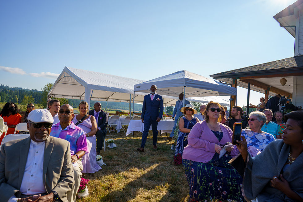 Guests seated and standing at sunny outdoor wedding ceremony.