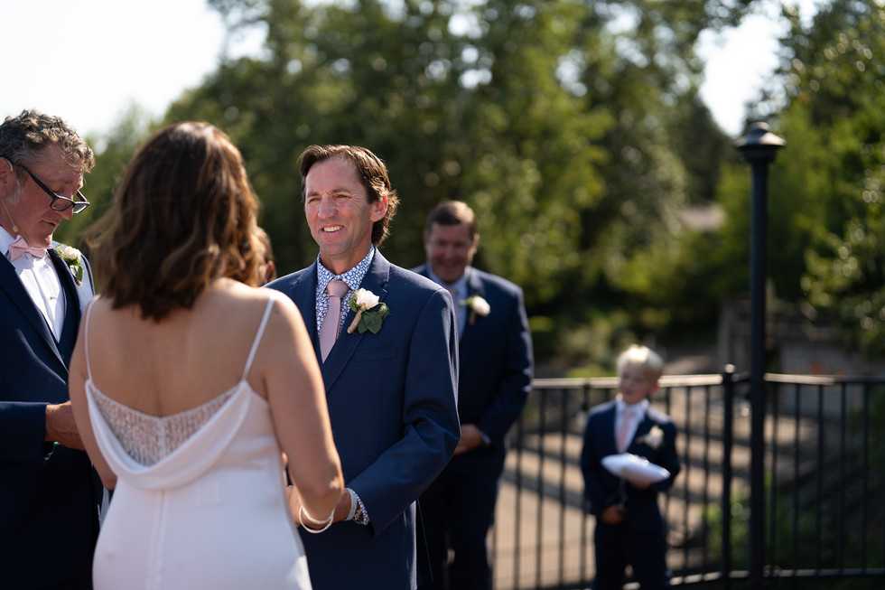 Groom in blue suit smiles at bride during sunny outdoor wedding.