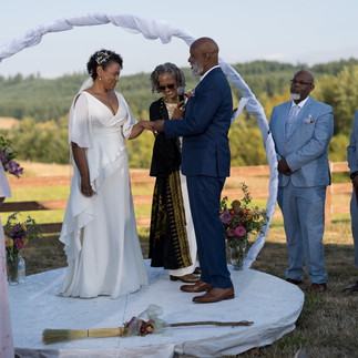 Black couple exchanging rings under arch during outdoor wedding ceremony.