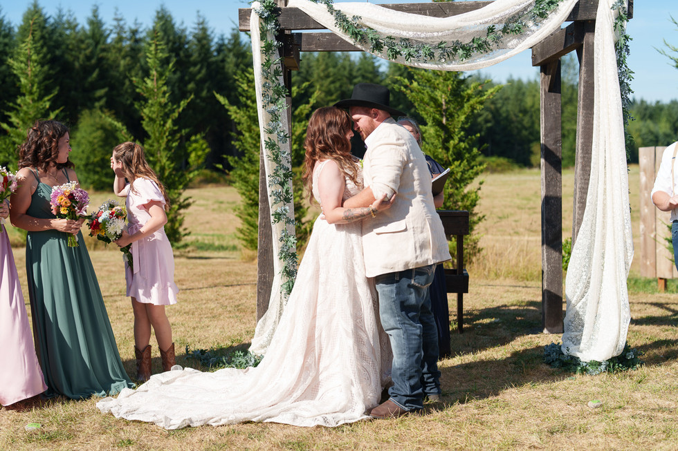 Bride and groom embrace under rustic arch at outdoor summer wedding.