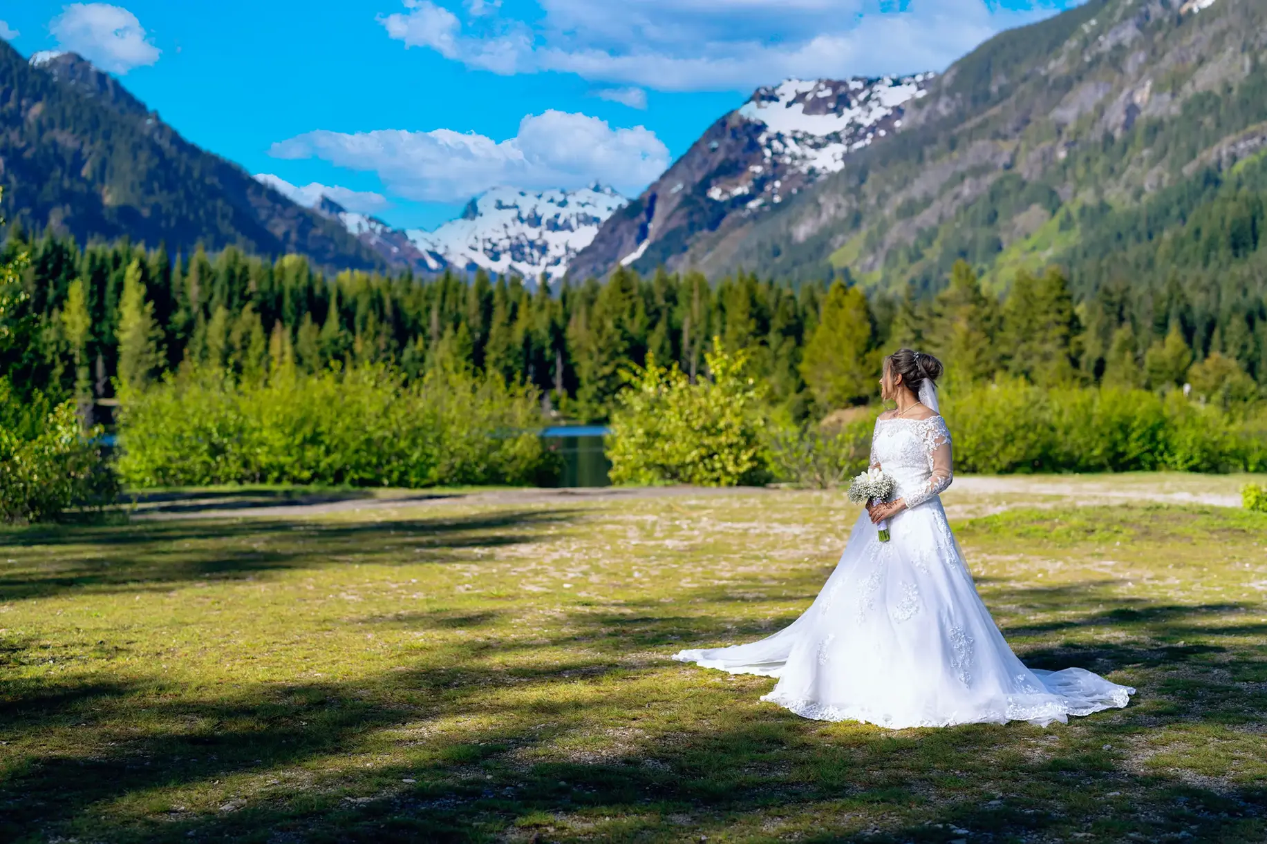 White-gowned bride, holding bouquet, snowy mountains, green forest, PNW wedding.