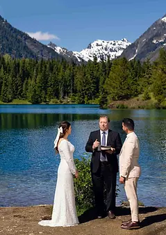 Couple exchanging vows during a wedding ceremony near a lake with mountains and trees. PhotoYari Photography