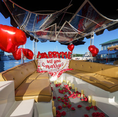 Romantic boat proposal: "Marry Me" banner, red heart balloons, rose petals
