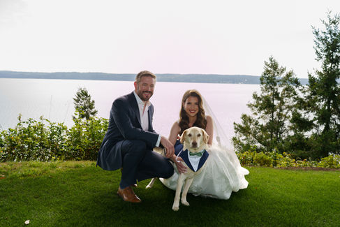 Bride, groom, and dog in tuxedo pose by lake for wedding.