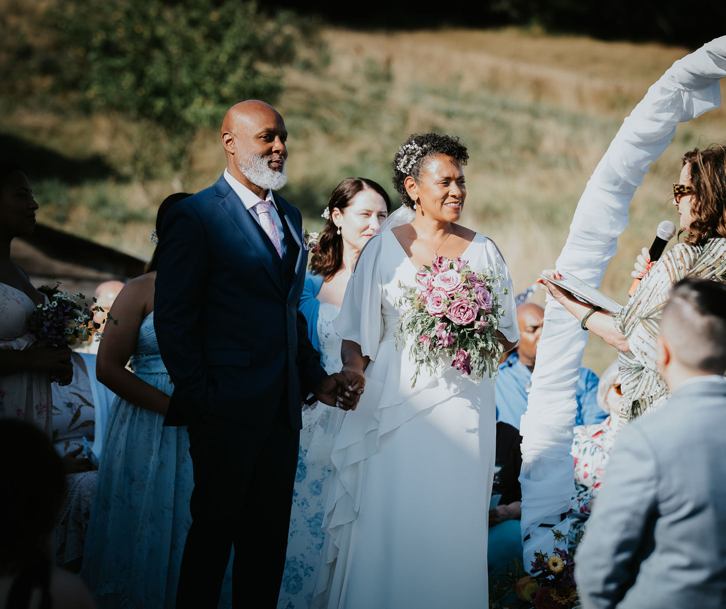 Diverse couple holding hands at outdoor wedding as officiant speaks.
