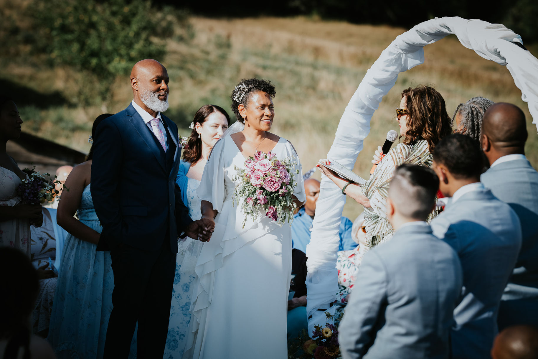 Diverse couple holding hands at outdoor wedding as officiant speaks.
